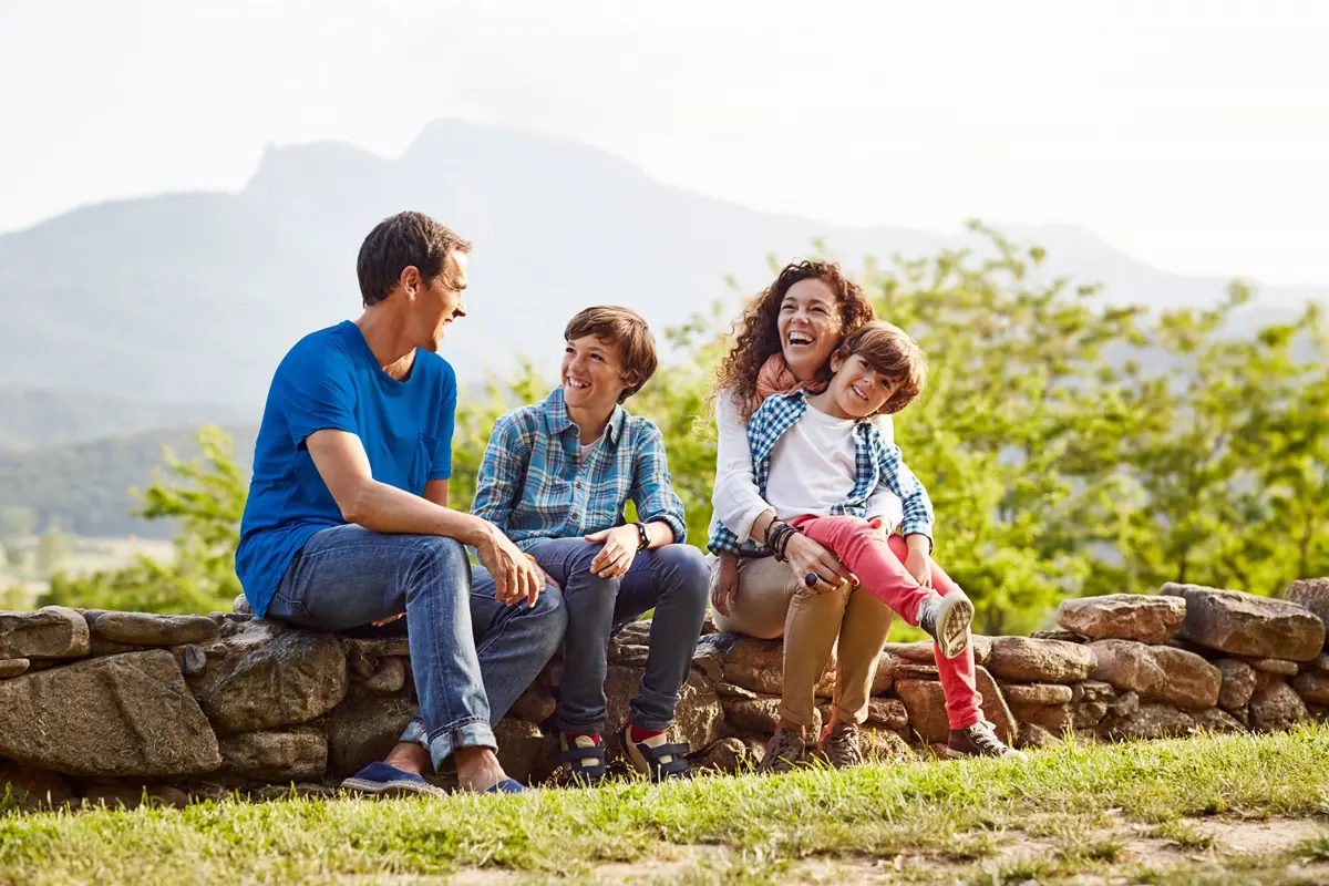 Familia de cuatro personas disfrutando al aire libre, sentada en un muro de piedra con montañas al fondo